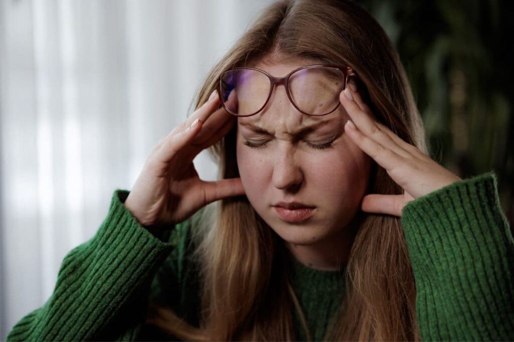 A woman with long hair holds her head in confusion, eyes closed, and wearing a green sweater. Glasses are pushed up on her forehead.