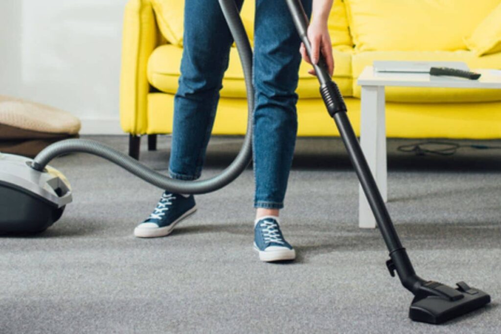 Woman vacuuming living room floor, yellow couch in background.