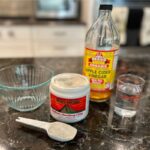 A kitchen counter displays a glass bowl, a jar of Bentonite Clay, a spoon with clay, a bottle of apple cider vinegar, and a glass of water—ingredients for an armpit detox using natural products to reduce odor and clear buildup.