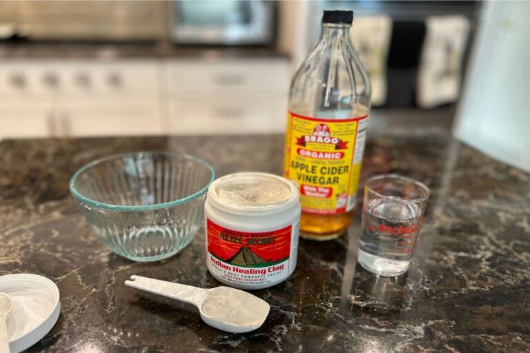 A kitchen counter displays a glass bowl, a jar of Bentonite Clay, a spoon with clay, a bottle of apple cider vinegar, and a glass of water—ingredients for an armpit detox using natural products to reduce odor and clear buildup.