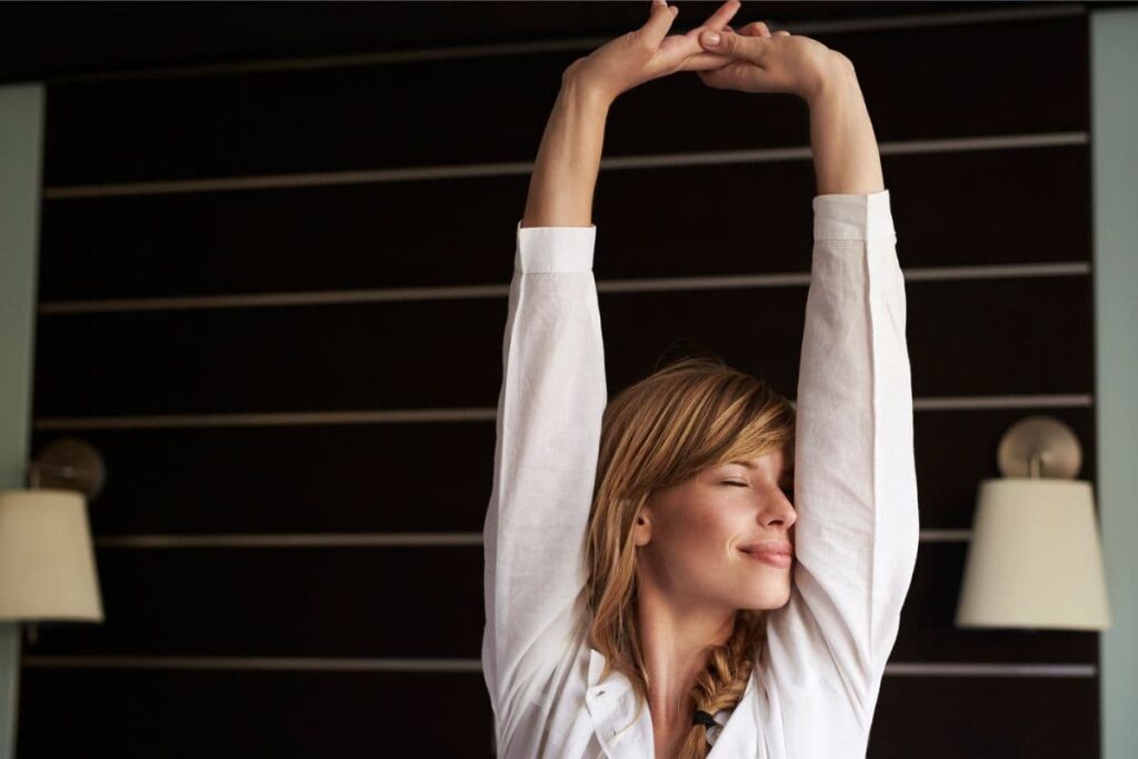 Woman in a white shirt stretches with eyes closed and nose in her armpit. She appears to be smelling her armpits and is smiling contentedly.