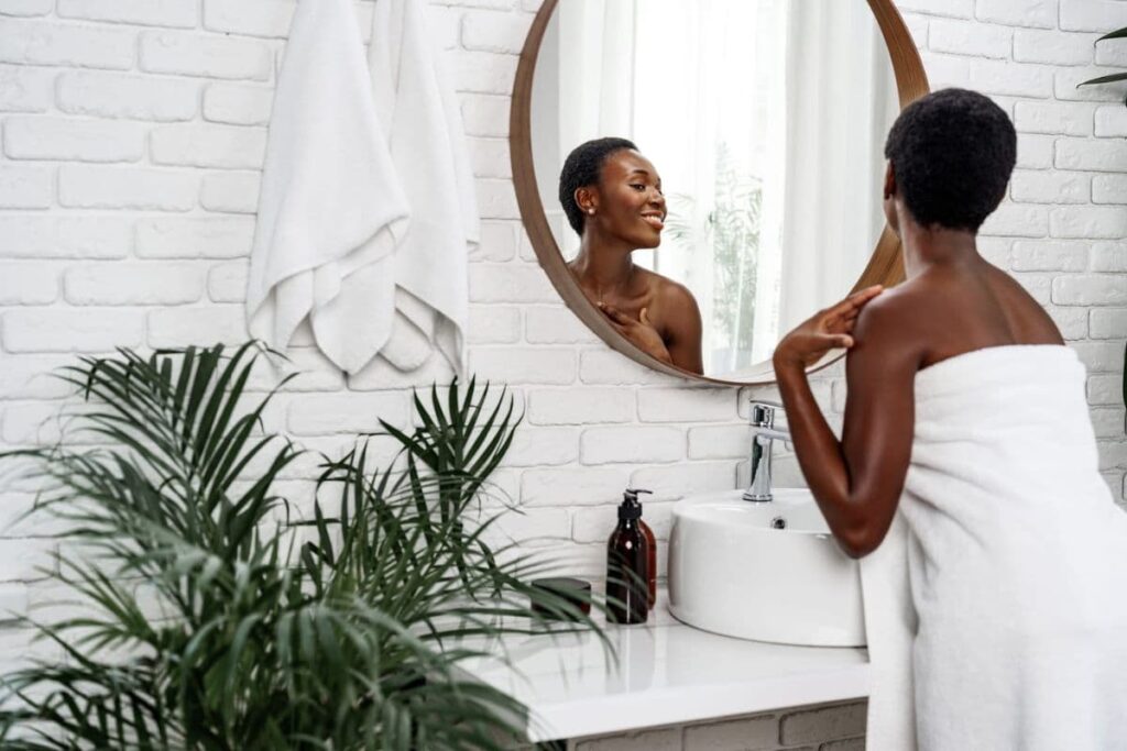 A woman wrapped in a towel smiles at her reflection in a round bathroom mirror.