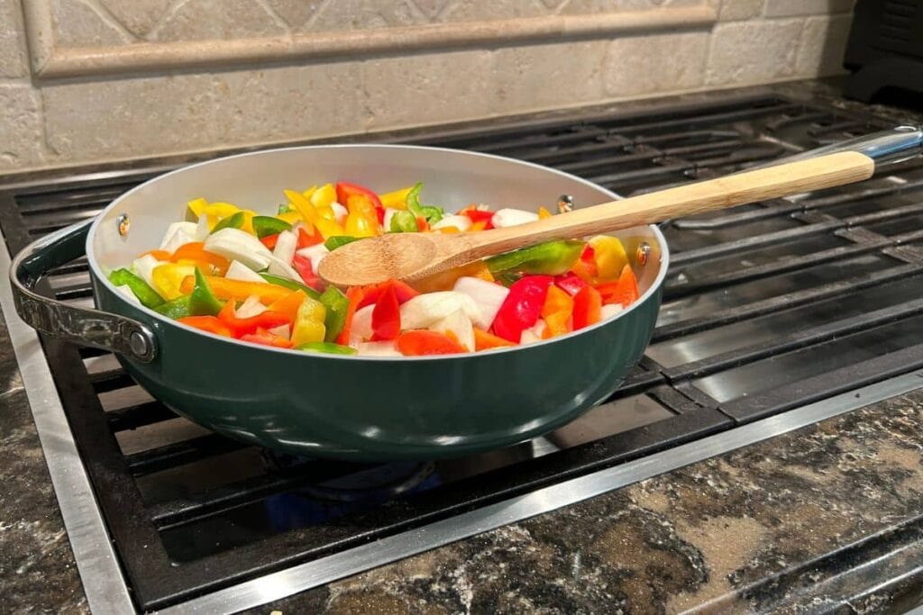 Colorful bell peppers and onions cooking in a sage green Caraway Home large skillet.