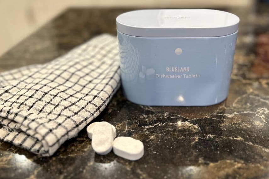 Blueland dishwasher tabs in forever tin on counter, three tabs and striped towel next to tin.