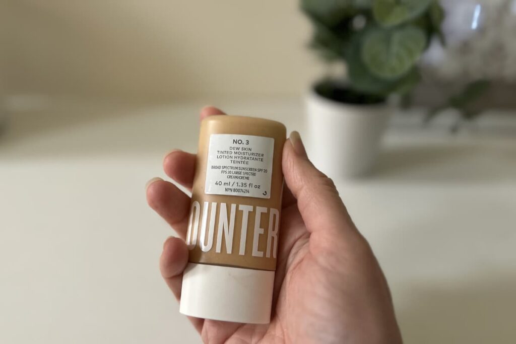 Hand holding Counter Dew Skin Tinted SPF Moisturizer, plant and picture out of focus in background