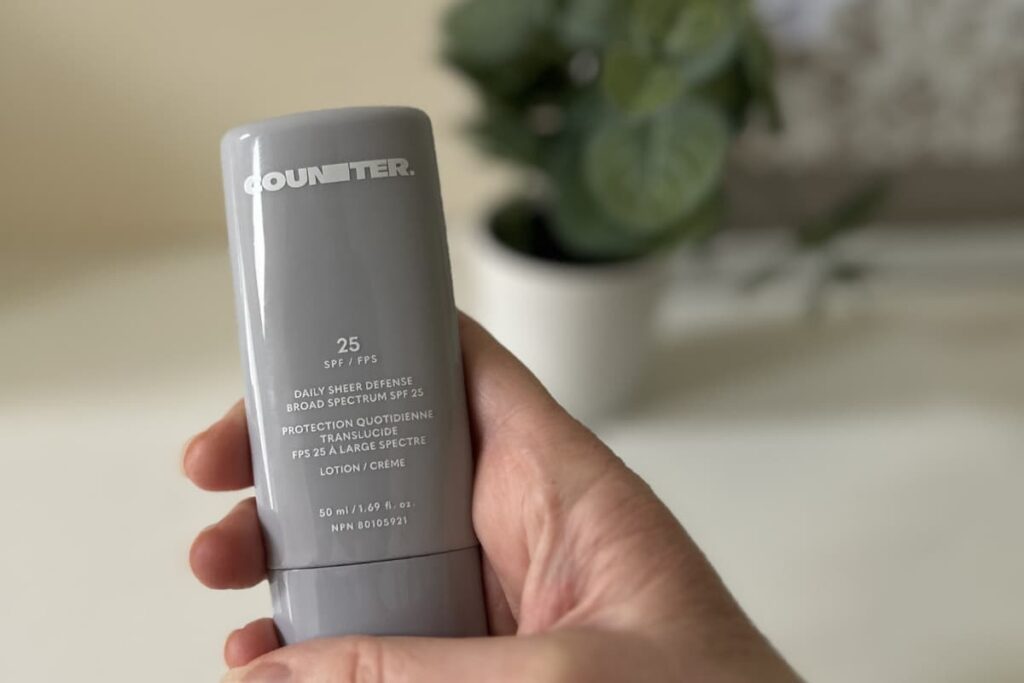 Hand holding Counter Daily Sheer Defense Sunscreen, plant and picture out of focus in background