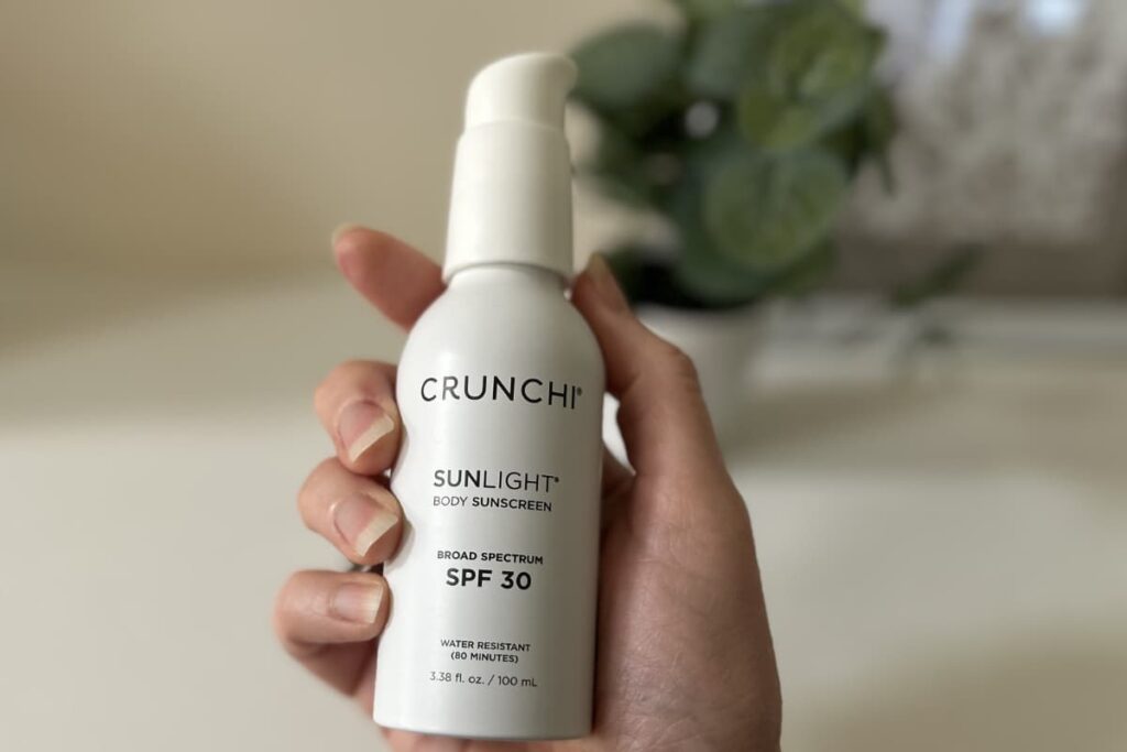 Hand holding Crunchi Sunlight Body Sunscreen, plant and picture out of focus in background