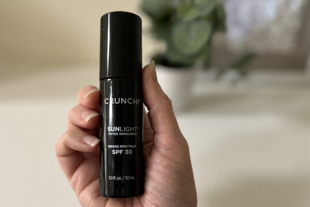 Hand holding Crunchi Tinted Sunscreen, plant and picture out of focus in background