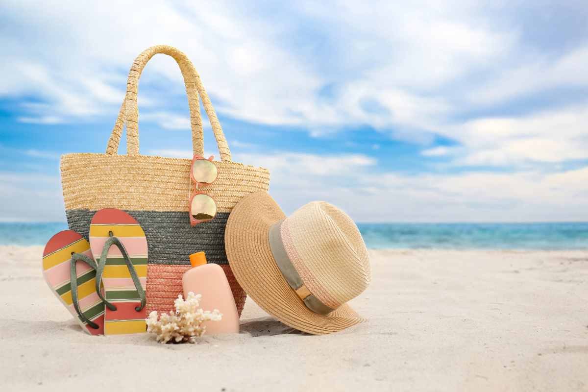 A straw bag, sunglasses, flip-flops, sunscreen, and hat on a sandy beach. Blue sky and ocean in the background.